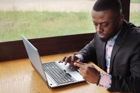 Black man makes payment bank card on laptop. Online shopping sitting in cafe. Young afro american businessman is buying online entering his data to computer from credit card, side view. Guy customer.の写真素材