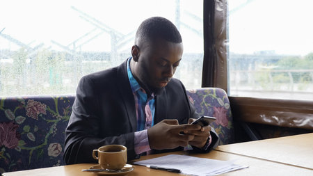 Afro american businessman is browsing smartphone sitting in cafe. He is typing a message chatting in...