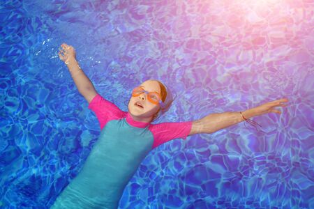 Portrait of calm girl relaxing in clear blue water of outdoors sunny swimming pool. Child in goggles, swimsuit and cap lying calmly on back.の写真素材