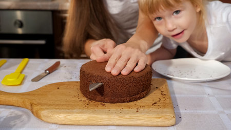 Family portrait of mom and daughters are preparing birthday cake together, mom is cutting sponge...