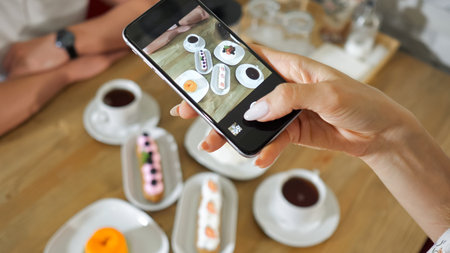young woman takes picture of different tasty cakes and tea cups with modern smartphone at wooden...