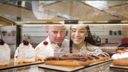 happy young couple chooses tasty fresh cakes standing in glass show case of confectionery shop close viewの写真素材