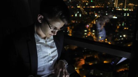 Young man freelancer in glasses is working on his computer near the window in home at night. Self-employed man has overtime job. Freelance and self-employed concept.の写真素材