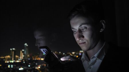 Young successful businessman in office suit is browsing mobile phone. He is standing near the window with panoramic city view in his office at night.の写真素材