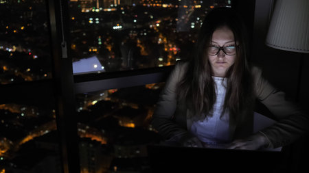 Brunette woman in glasses is working on her computer near the window in home at night. Young...