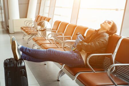 Young woman sleeping with her legs stretched out on the suitcase on the chairs at the waiting room of the airport, sunlight. Long waiting for the airplane.の写真素材