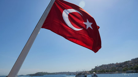 red turkish flag flatters against distant cityscape and ships sailing in sea sparkling under bright...