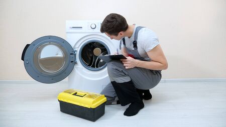 skilled repairman writes on clipboard checking broken washing machine on floor in room closeupの写真素材