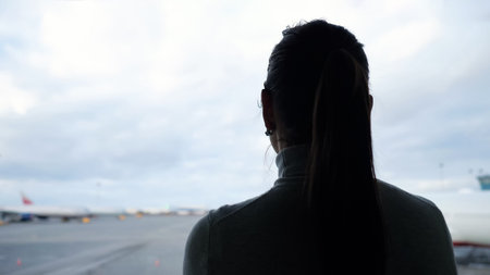 woman silhouette with ponytail looks at airplanes on airfield apron from airport terminal close...