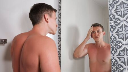 handsome guy with bare torso looks at sunburned face and body in mirror standing in hotel bathroom close viewの写真素材
