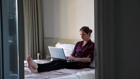 attractive dark haired lady sits on white bed and types on grey modern laptop with serious face at home in morningの写真素材