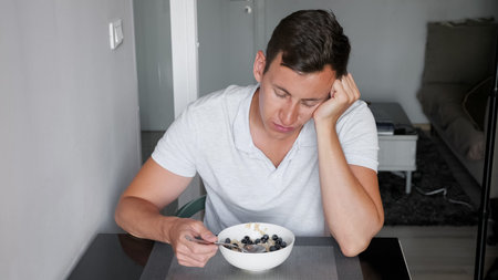 sleepy sunburned man in t-shirt sits at table resting head with hand and eats healthy breakfast at...