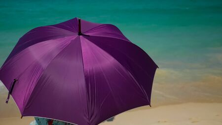 woman in dress with flower pattern sits on yellow beach holding purple umbrella hiding from sun against ocean backside viewの写真素材