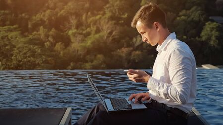 young man in white shirt works on laptop taking look at phone against green plants and blue pool water at sunset lightの写真素材