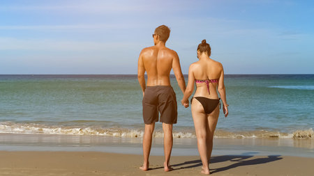 pretty young couple walk along beach to ocean water joining hands at tropical resort backside view...