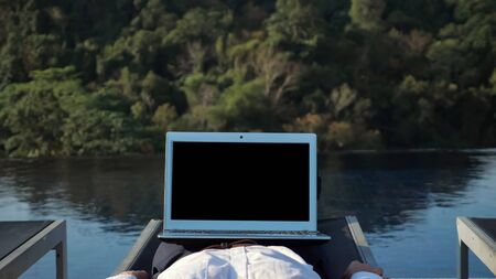 laptop with dark screen on legs of relaxing in lounge chair man against green forestry hill casting shadow on blue poolの写真素材