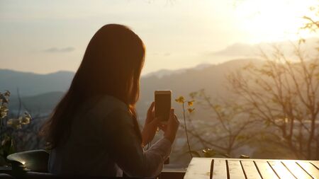 Young woman photographs sunset on the phone in a cafe on a hill copyspaceの写真素材