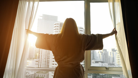 girl silhouette in bathrobe opens window transparent curtains looking at buildings outside in hotel...