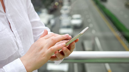 lady in white blouse stands on road bridge holding modern smartphone and types against blurry cars...