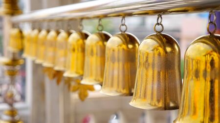 beautiful golden traditional Buddhist bells waved by wind hang on long silver pipe against blurry local buildings closeupの写真素材