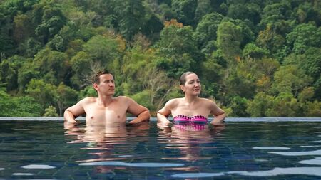 smiling couple relaxes in large hotel swimming pool blue water against green hilly landscape under bright summer sunlightの写真素材