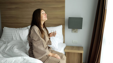 happy brunette lady in beige bathrobe drinks clean water against bright window in comfortable hotel...