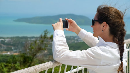 brunette in white blouse and sunglasses takes photo of marvelous cityscape and endless ocean with...