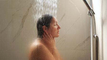 attractive brunette stands under heavy water pressure and washes face in home bathroom shower cabin...