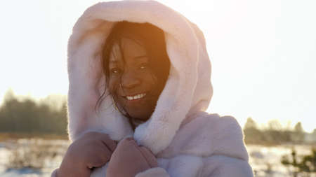 positive African-American lady in white fur hood poses smiling against empty forest in cold winter at back sunlight closeupの写真素材