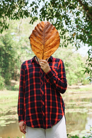 Young woman in a checkered shirt in black and red covers her face with a large, dried leaf of a...