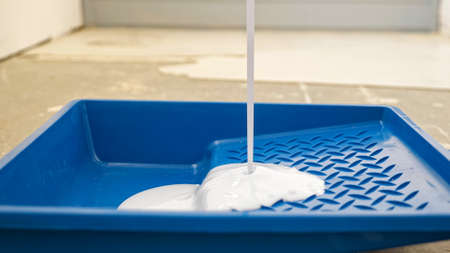 pouring white acrylic paint into blue plastic tray to make improvement on floor in spacious brightly lit room extreme close viewの写真素材