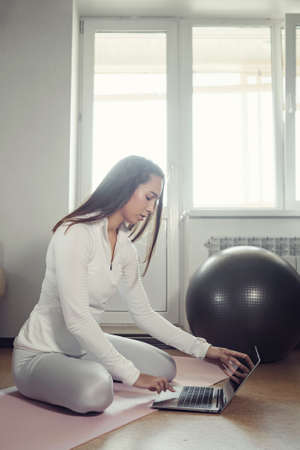 pretty woman in white tracksuit surfs internet with modern laptop to find new exercises on mat at homeの写真素材