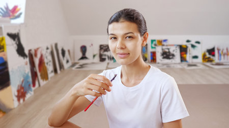 young pretty woman with brush poses smiling against various own paintings on brown wooden floor...