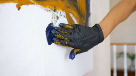 Female hand in black rubber glove runs on white canvas drawing blue waves in atelier with white walls under bright light close-upの写真素材