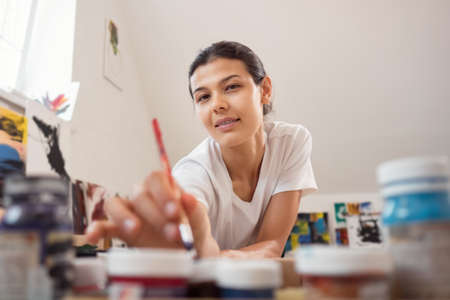 Beautiful young woman in a white t-shirt with a brush in her hand against a background of blurry paintings.の写真素材