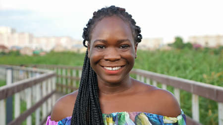 attractive African-American girl with long cornrows in bright color blouse smiles standing on wooden bridge in green reeds closeupの写真素材