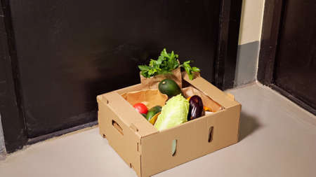 Skilled delivery guy in denim jeans puts brown cardboard container with fresh vegetables on floor near black door closeupの写真素材