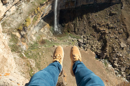 Person in blue jeans and beige sneakers sits swinging legs on high cliff edge near narrow waterfall...