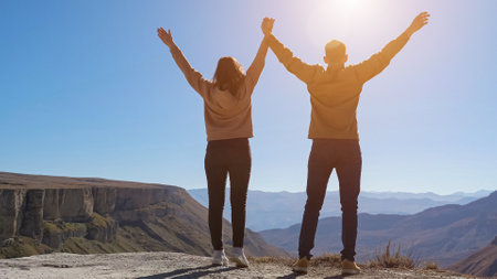Young couple walks and raises joined hands enjoying view from cliff edge against distant mountains and blue sky on sunny day backside viewの写真素材