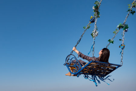 Barefoot brunette in long polka dot dress sits on large designed swings decorated with green leaves and coloured tapes under blue skyの写真素材