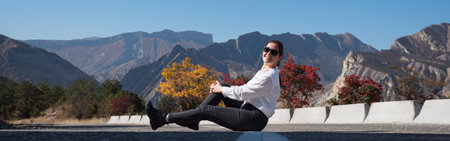 Young woman in white hoodie and sunglasses sits on grey asphalt road barrier against red and orange...