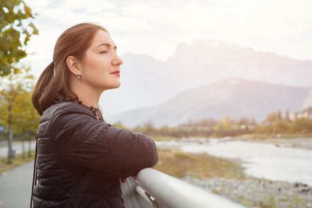 Young woman with long loose flowing hair stands near grey metal handrails and looks at river against tree silhouettes lit by bright sunの写真素材