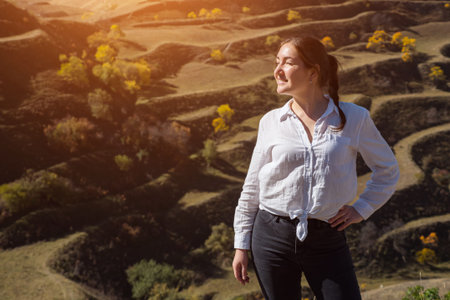 Brunette in white blouse and black jeans poses for camera standing against terraced hill with fields...