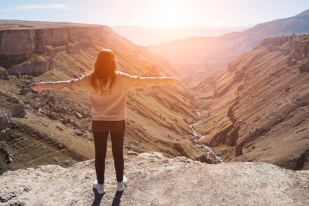 Long haired brunette in sportswear stands on rocky hill edge and admires mountainous landscape lit by bright autumn sunlight backside viewの写真素材