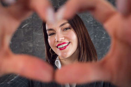 Young brunette with short hair and red lipstick shows heart sign with fingers and smiles standing against grey office wall closeupの写真素材