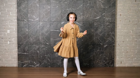 Delighted active girl in brown dress dances wearing black headphones against designed marble wall...