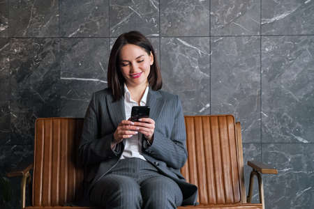 Businesswoman with short dark hair types on black smartphone sitting on brown bench against grey and white marble wall in new office receptionの写真素材