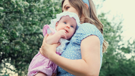Careful blonde mother in blue dress holds in arms crying girl kid and lulls standing against blurred blooming fruit trees in park on spring dayの写真素材