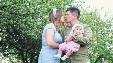 Young father in green shirt holding baby girl kisses mother wearing summer dress in city park against blossoming fruit trees with green leavesの写真素材