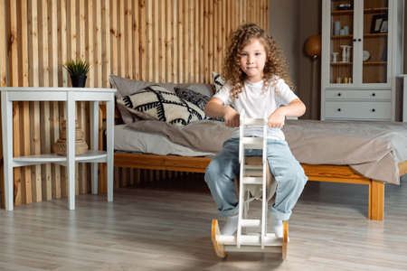 Cute preschooler girl with long loose curly brown hair sits on white wooden rocking horse and rocks against stylish furniture in bedroomの写真素材